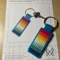 A hand holds a finished needlepoint keychain and a set of keys. The keychain is rectangular with a striped pattern in shades of blue, green, yellow, pink, and red, and is attached to a silver key ring.
