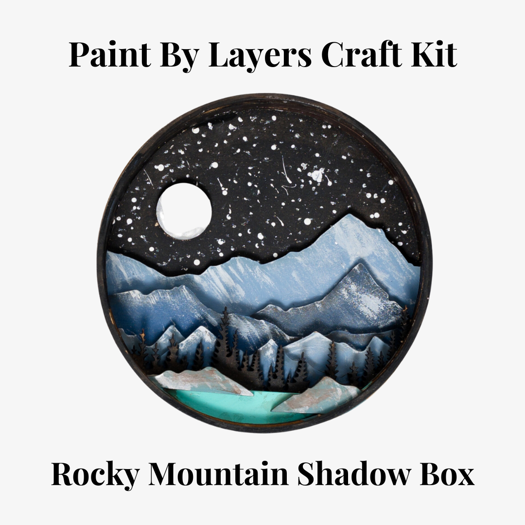 A Paint By Layers Craft Kit featuring a circular shadow box with a pre-designed landscape scene. The scene depicts a mountain range under a starry night sky, with a river flowing through a valley. The kit includes layers of wood, paint, and instructions. The title of the kit is "Rocky Mountain Shadow Box."