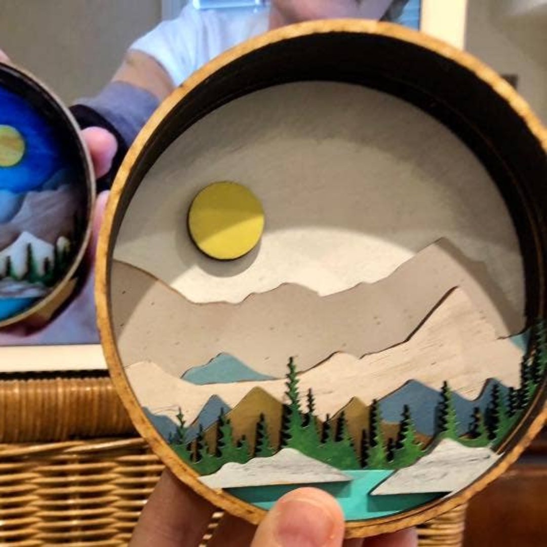 A Paint By Layers Craft Kit featuring a circular shadow box with a pre-designed landscape scene. The scene depicts a mountain range under a starry night sky, with a river flowing through a valley. The kit includes layers of wood, paint, and instructions. The title of the kit is "Rocky Mountain Shadow Box."