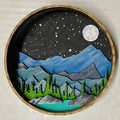 A Paint By Layers Craft Kit featuring a circular shadow box with a pre-designed landscape scene. The scene depicts a mountain range under a starry night sky, with a river flowing through a valley. The kit includes layers of wood, paint, and instructions. The title of the kit is "Rocky Mountain Shadow Box."