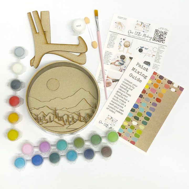 A Paint By Layers Craft Kit featuring a circular shadow box with a pre-designed landscape scene. The scene depicts a mountain range under a starry night sky, with a river flowing through a valley. The kit includes layers of wood, paint, and instructions. The title of the kit is 