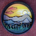 A Paint By Layers Craft Kit featuring a circular shadow box with a pre-designed landscape scene. The scene depicts a mountain range under a starry night sky, with a river flowing through a valley. The kit includes layers of wood, paint, and instructions. The title of the kit is "Rocky Mountain Shadow Box."