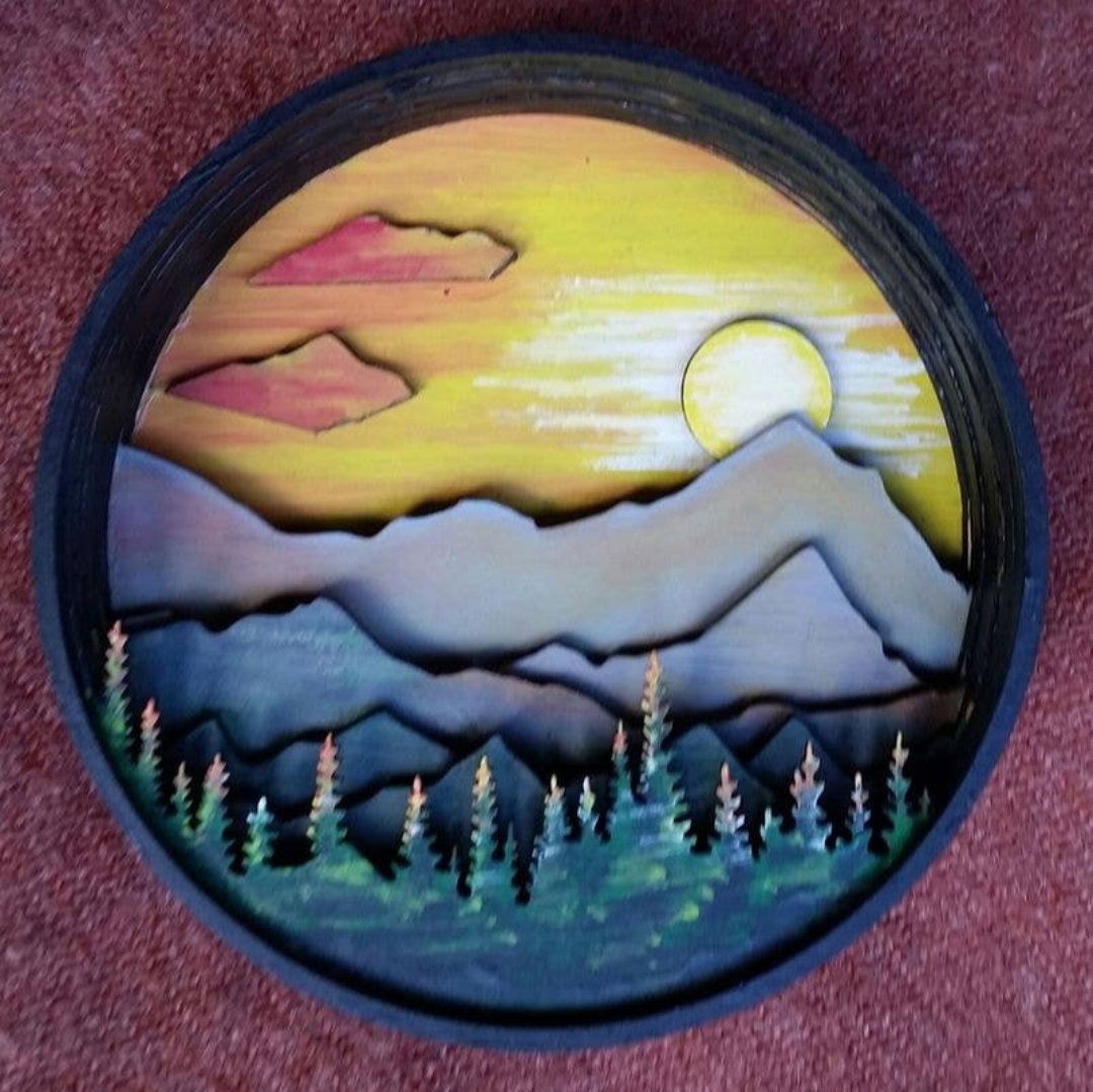 A Paint By Layers Craft Kit featuring a circular shadow box with a pre-designed landscape scene. The scene depicts a mountain range under a starry night sky, with a river flowing through a valley. The kit includes layers of wood, paint, and instructions. The title of the kit is "Rocky Mountain Shadow Box."