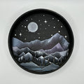 A Paint By Layers Craft Kit featuring a circular shadow box with a pre-designed landscape scene. The scene depicts a mountain range under a starry night sky, with a river flowing through a valley. The kit includes layers of wood, paint, and instructions. The title of the kit is "Rocky Mountain Shadow Box."
