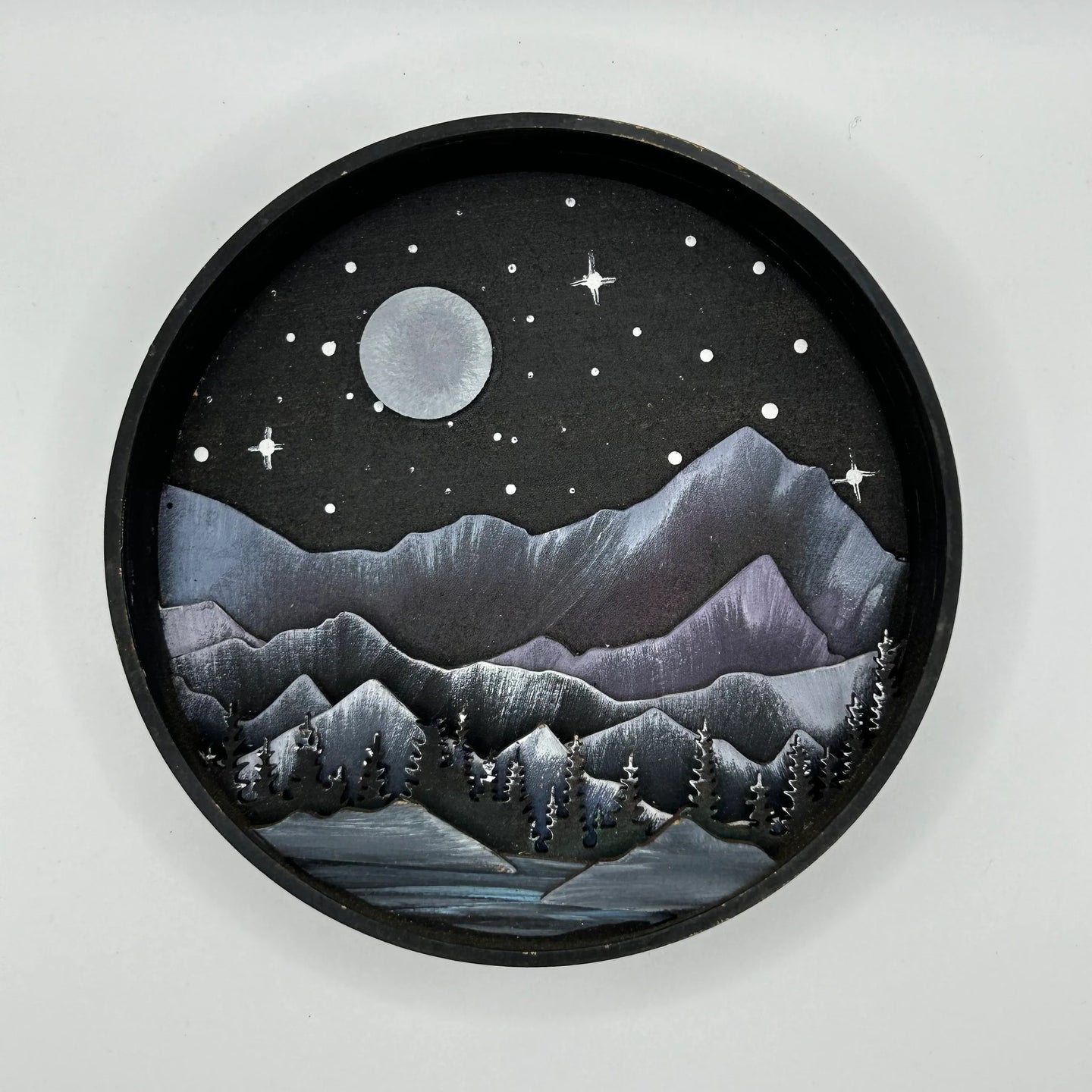 A Paint By Layers Craft Kit featuring a circular shadow box with a pre-designed landscape scene. The scene depicts a mountain range under a starry night sky, with a river flowing through a valley. The kit includes layers of wood, paint, and instructions. The title of the kit is "Rocky Mountain Shadow Box."