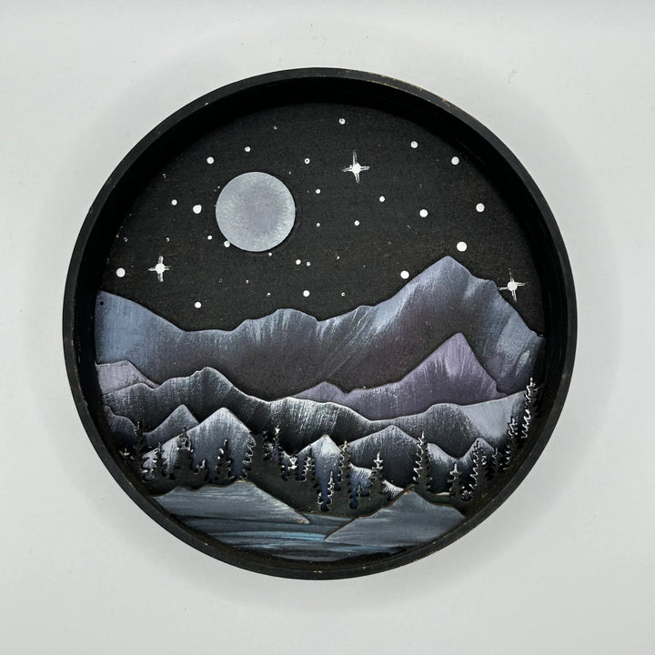 A Paint By Layers Craft Kit featuring a circular shadow box with a pre-designed landscape scene. The scene depicts a mountain range under a starry night sky, with a river flowing through a valley. The kit includes layers of wood, paint, and instructions. The title of the kit is 