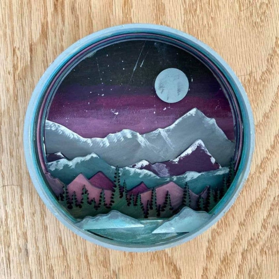 A Paint By Layers Craft Kit featuring a circular shadow box with a pre-designed landscape scene. The scene depicts a mountain range under a starry night sky, with a river flowing through a valley. The kit includes layers of wood, paint, and instructions. The title of the kit is "Rocky Mountain Shadow Box."