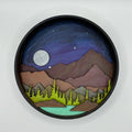 A Paint By Layers Craft Kit featuring a circular shadow box with a pre-designed landscape scene. The scene depicts a mountain range under a starry night sky, with a river flowing through a valley. The kit includes layers of wood, paint, and instructions. The title of the kit is "Rocky Mountain Shadow Box."