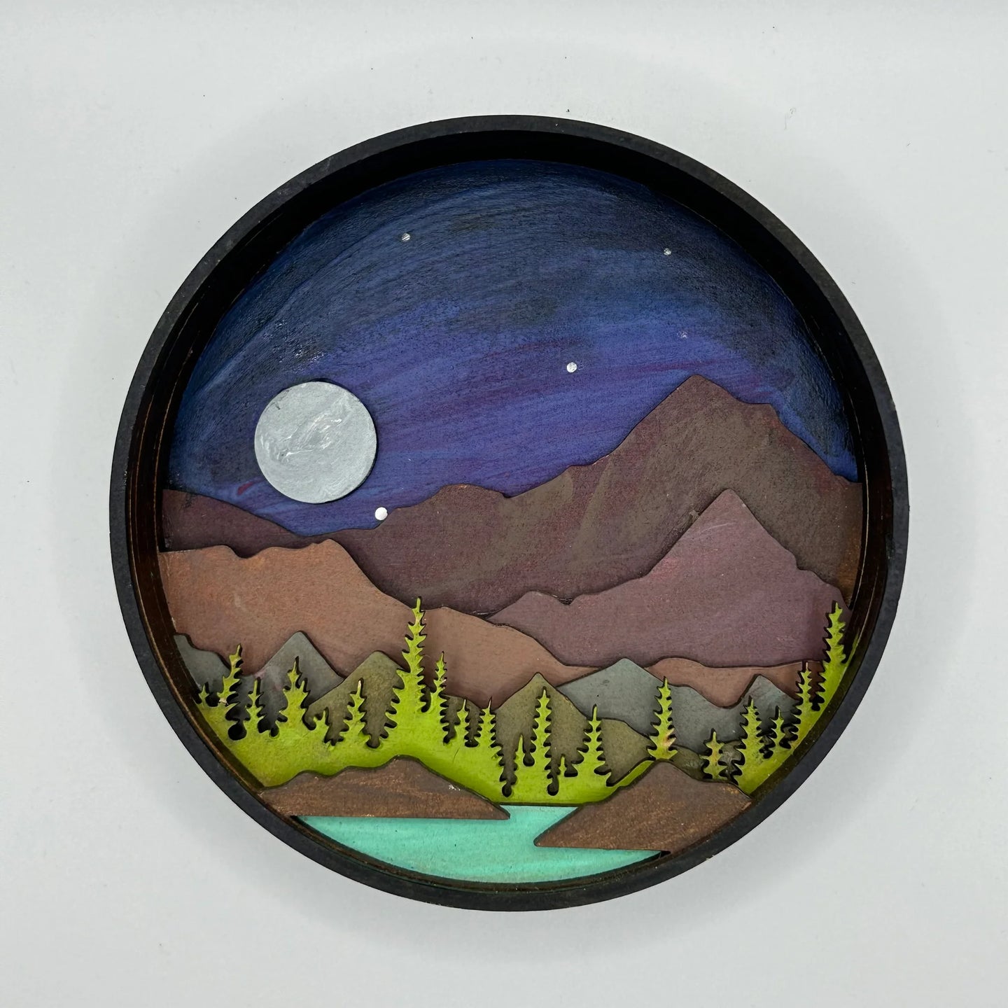 A Paint By Layers Craft Kit featuring a circular shadow box with a pre-designed landscape scene. The scene depicts a mountain range under a starry night sky, with a river flowing through a valley. The kit includes layers of wood, paint, and instructions. The title of the kit is "Rocky Mountain Shadow Box."