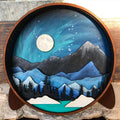 A Paint By Layers Craft Kit featuring a circular shadow box with a pre-designed landscape scene. The scene depicts a mountain range under a starry night sky, with a river flowing through a valley. The kit includes layers of wood, paint, and instructions. The title of the kit is "Rocky Mountain Shadow Box."