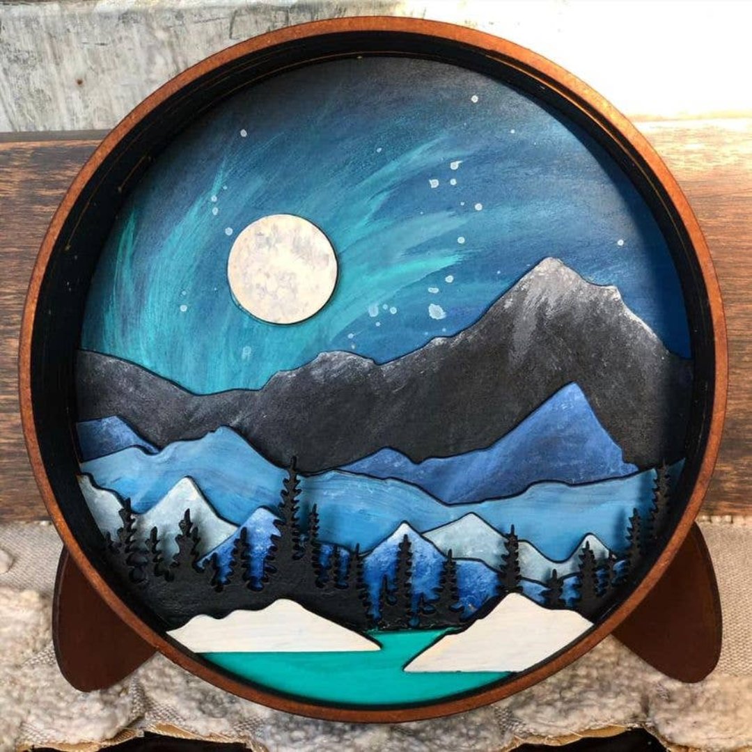 A Paint By Layers Craft Kit featuring a circular shadow box with a pre-designed landscape scene. The scene depicts a mountain range under a starry night sky, with a river flowing through a valley. The kit includes layers of wood, paint, and instructions. The title of the kit is "Rocky Mountain Shadow Box."