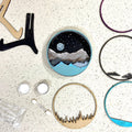 A Paint By Layers Craft Kit featuring a circular shadow box with a pre-designed landscape scene. The scene depicts a mountain range under a starry night sky, with a river flowing through a valley. The kit includes layers of wood, paint, and instructions. The title of the kit is "Rocky Mountain Shadow Box."