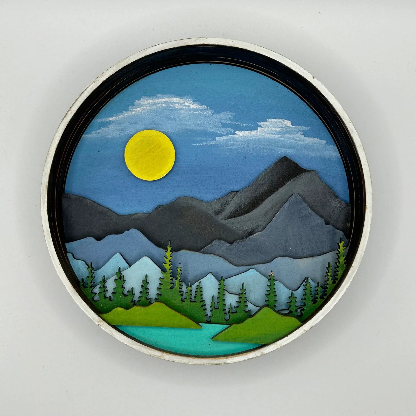 A Paint By Layers Craft Kit featuring a circular shadow box with a pre-designed landscape scene. The scene depicts a mountain range under a starry night sky, with a river flowing through a valley. The kit includes layers of wood, paint, and instructions. The title of the kit is "Rocky Mountain Shadow Box."