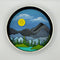 A Paint By Layers Craft Kit featuring a circular shadow box with a pre-designed landscape scene. The scene depicts a mountain range under a starry night sky, with a river flowing through a valley. The kit includes layers of wood, paint, and instructions. The title of the kit is 