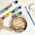 A Paint By Layers Craft Kit featuring a circular shadow box with a pre-designed landscape scene. The scene depicts a mountain range under a starry night sky, with a river flowing through a valley. The kit includes layers of wood, paint, and instructions. The title of the kit is "Rocky Mountain Shadow Box."