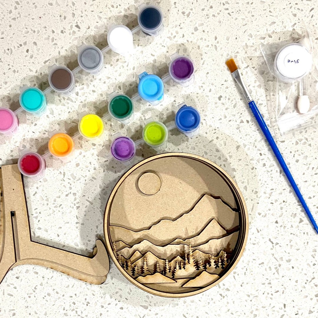 A Paint By Layers Craft Kit featuring a circular shadow box with a pre-designed landscape scene. The scene depicts a mountain range under a starry night sky, with a river flowing through a valley. The kit includes layers of wood, paint, and instructions. The title of the kit is "Rocky Mountain Shadow Box."