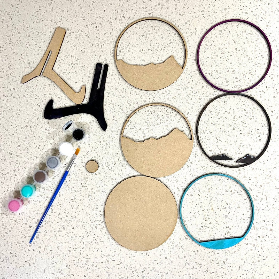 A Paint By Layers Craft Kit featuring a circular shadow box with a pre-designed landscape scene. The scene depicts a mountain range under a starry night sky, with a river flowing through a valley. The kit includes layers of wood, paint, and instructions. The title of the kit is "Rocky Mountain Shadow Box."