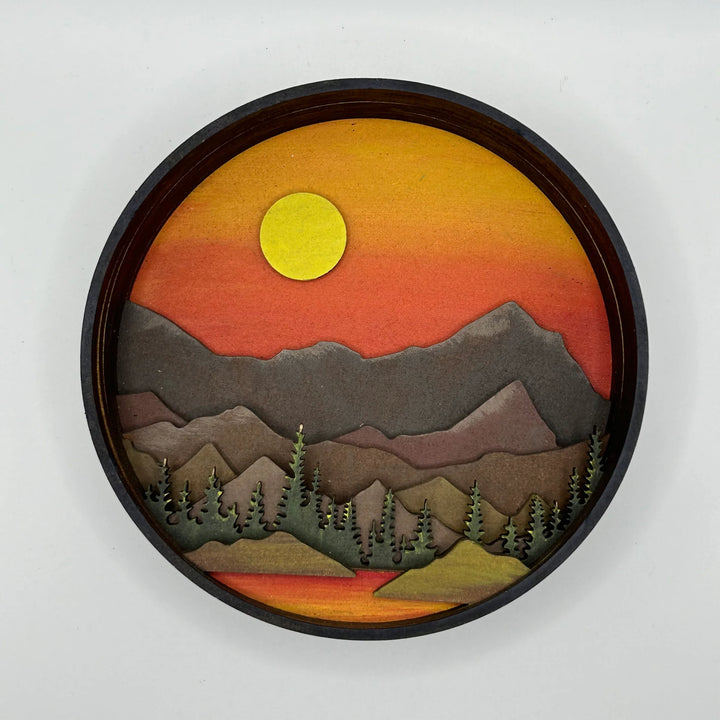 A Paint By Layers Craft Kit featuring a circular shadow box with a pre-designed landscape scene. The scene depicts a mountain range under a starry night sky, with a river flowing through a valley. The kit includes layers of wood, paint, and instructions. The title of the kit is 