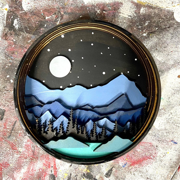 A Paint By Layers Craft Kit featuring a circular shadow box with a pre-designed landscape scene. The scene depicts a mountain range under a starry night sky, with a river flowing through a valley. The kit includes layers of wood, paint, and instructions. The title of the kit is 