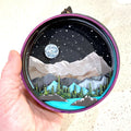 A Paint By Layers Craft Kit featuring a circular shadow box with a pre-designed landscape scene. The scene depicts a mountain range under a starry night sky, with a river flowing through a valley. The kit includes layers of wood, paint, and instructions. The title of the kit is "Rocky Mountain Shadow Box."