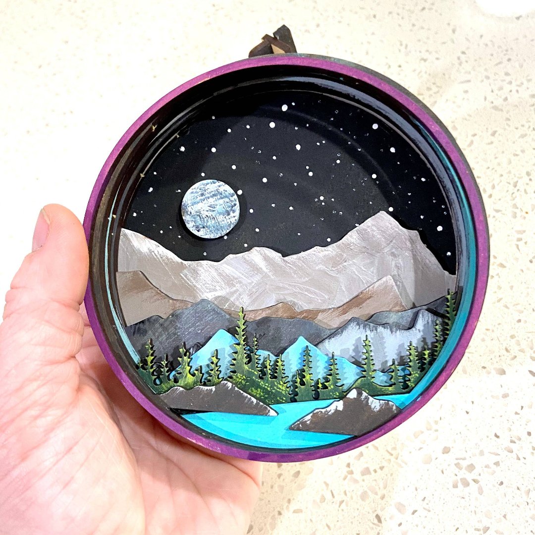 A Paint By Layers Craft Kit featuring a circular shadow box with a pre-designed landscape scene. The scene depicts a mountain range under a starry night sky, with a river flowing through a valley. The kit includes layers of wood, paint, and instructions. The title of the kit is "Rocky Mountain Shadow Box."