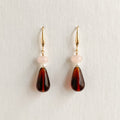 A pair of drop earrings with gold-tone hooks, a small pale pink round bead, a tiny white pearl, and a larger teardrop-shaped dark brown marbled bead, displayed against a light off-white background.