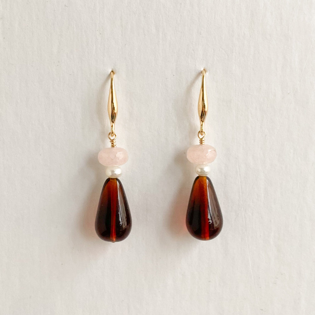 A pair of drop earrings with gold-tone hooks, a small pale pink round bead, a tiny white pearl, and a larger teardrop-shaped dark brown marbled bead, displayed against a light off-white background.