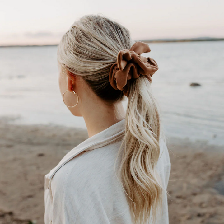 A rust-colored fabric scrunchie with a gathered, ruffled appearance. It has a small white tag with the word 