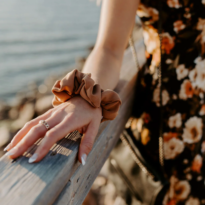A rust-colored fabric scrunchie with a gathered, ruffled appearance. It has a small white tag with the word 