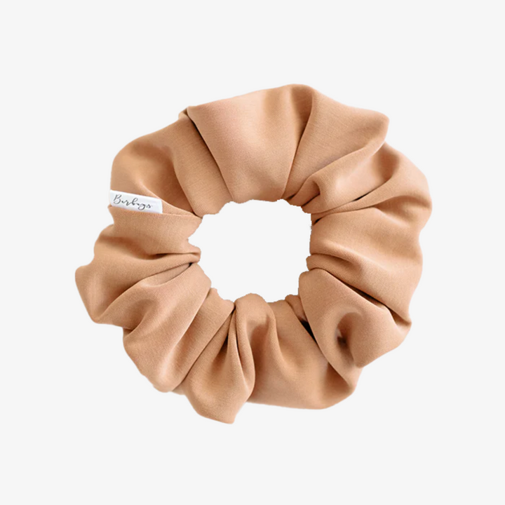 A rust-colored fabric scrunchie with a gathered, ruffled appearance. It has a small white tag with the word 