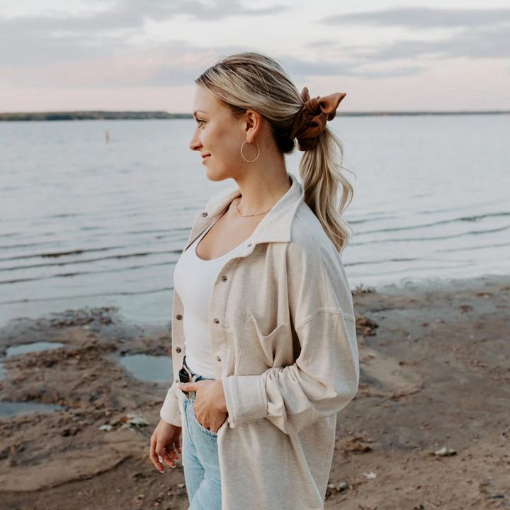 A rust-colored fabric scrunchie with a gathered, ruffled appearance. It has a small white tag with the word 