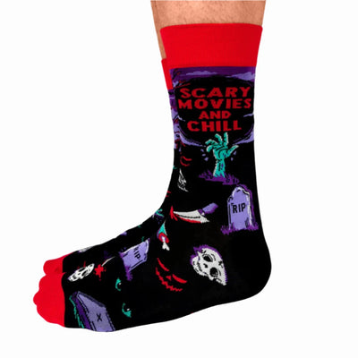 A pair of crew socks with a Halloween theme. The socks are mostly black with red and purple accents. The design includes a zombie hand, a graveyard with tombstones, a skull, and bats. The text 