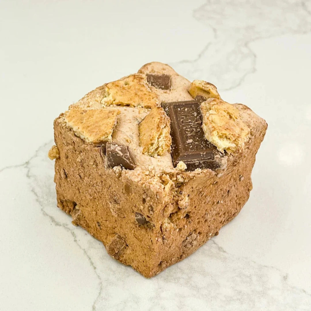 A square marshmallow with graham cracker crumbs, chocolate chunks, and melted marshmallow on top. The marshmallow is resting on a white marble surface.