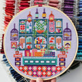 An overhead shot of a finished cross-stitch piece in a wooden embroidery hoop, displayed on a white surface with a potted succulent in the top right corner. The cross-stitch design features a colourful and intricate illustration of a shop interior with shelves, books, plants, and decorative items.