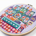 An overhead shot of a finished cross-stitch piece in a wooden embroidery hoop, displayed on a white surface with a potted succulent in the top right corner. The cross-stitch design features a colourful and intricate illustration of a shop interior with shelves, books, plants, and decorative items.