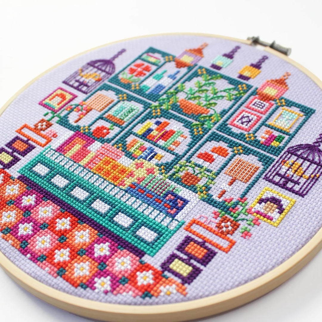 An overhead shot of a finished cross-stitch piece in a wooden embroidery hoop, displayed on a white surface with a potted succulent in the top right corner. The cross-stitch design features a colourful and intricate illustration of a shop interior with shelves, books, plants, and decorative items.