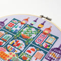 An overhead shot of a finished cross-stitch piece in a wooden embroidery hoop, displayed on a white surface with a potted succulent in the top right corner. The cross-stitch design features a colourful and intricate illustration of a shop interior with shelves, books, plants, and decorative items.