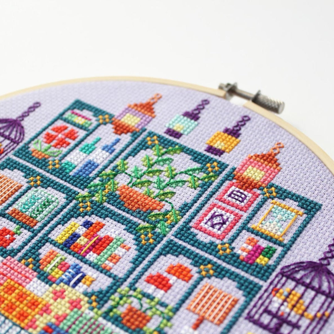 An overhead shot of a finished cross-stitch piece in a wooden embroidery hoop, displayed on a white surface with a potted succulent in the top right corner. The cross-stitch design features a colourful and intricate illustration of a shop interior with shelves, books, plants, and decorative items.