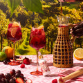 A styled outdoor picnic scene with two glasses of red fruit punch, a woven pitcher being filled, and a small bottle labeled 'Fruit Punch' on a pink tablecloth, set against a vineyard backdrop. A hand is visible holding the pitcher.