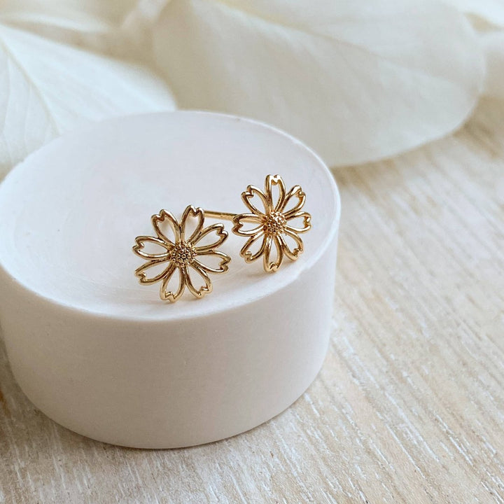A pair of gold daisy stud earrings on a white background. The earrings have a delicate, openwork design with a small circle in the center of each flower.