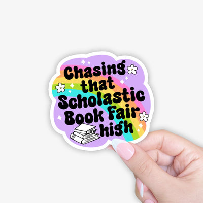 Sticker with 'Chasing that Scholastic Book Fair high' text on a colorful background held by a hand.