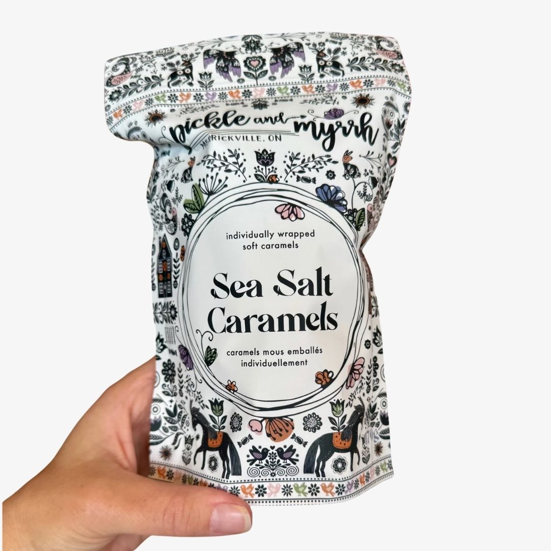 A hand holding a stand-up pouch of Pickle and Myrv Sea Salt Caramels against a plain white background. The packaging is white with black text and an intricate botanical and animal illustration pattern in muted tones of blue, orange, and green.