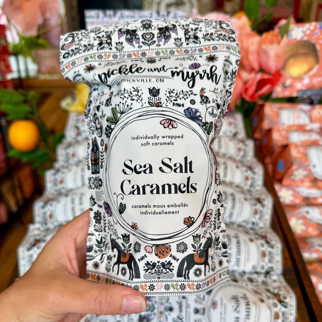 A hand holds a bag of Pickle and Myrk Sea Salt Caramels in front of a display of packaged caramels in a wooden tray.