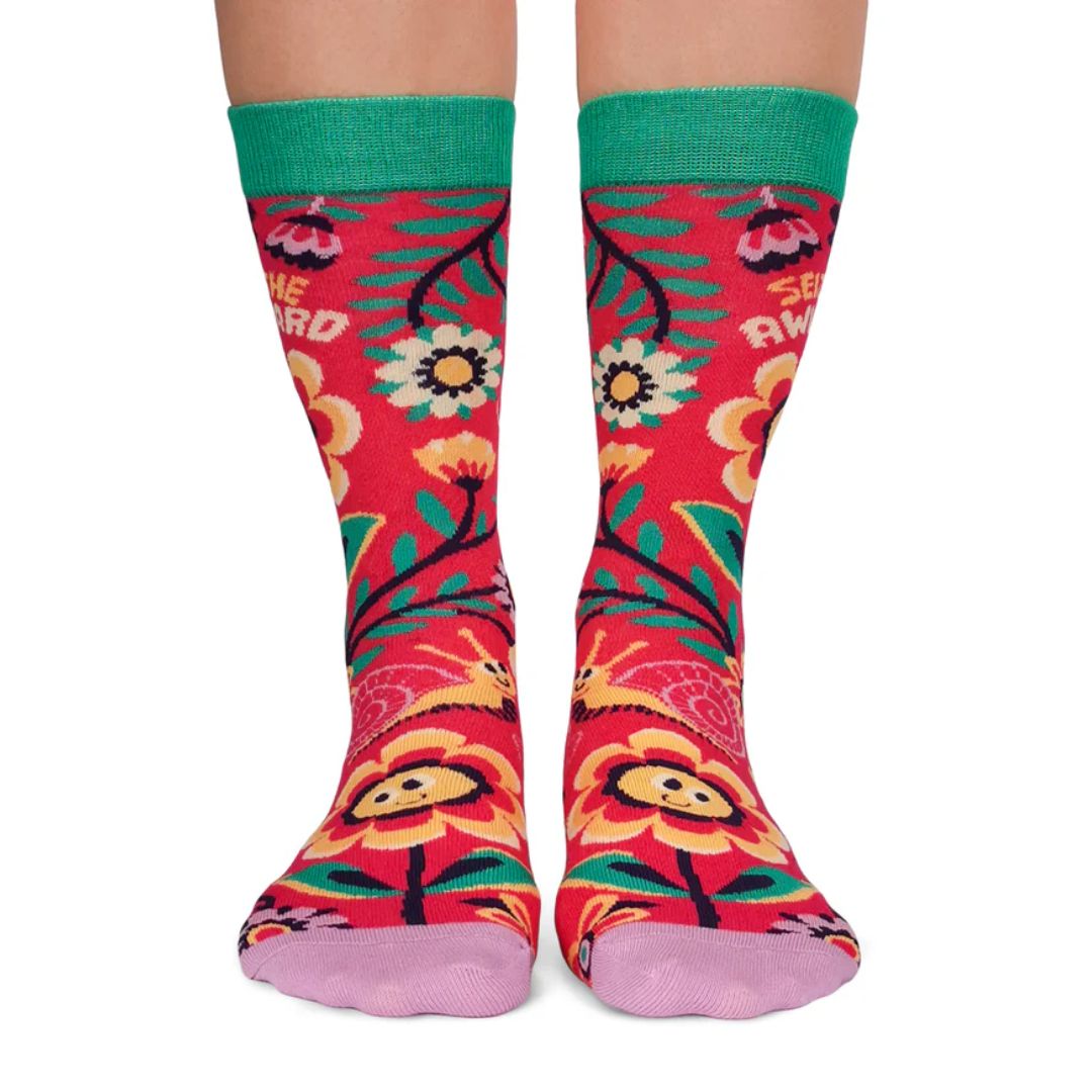 A pair of colourful women's crew socks with a red background featuring a whimsical floral pattern in shades of yellow, pink, green, and white. One sock displays the text "SEIZE THE AWKWARD" in white letters around a smiling yellow flower. The toe of the sock is light purple, the heel is yellow, and the cuff is bright green.