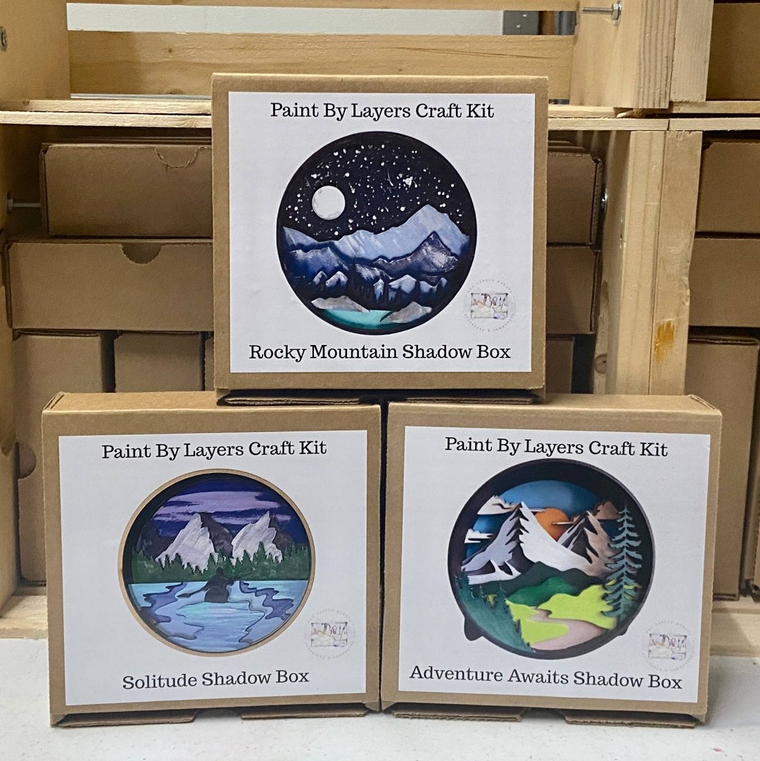 Three cardboard boxes containing Paint By Layers Craft Kits. The boxes are stacked on top of each other. The front of each box shows a circular image of the finished product. The titles of the kits are "Rocky Mountain Shadow Box," "Solitude Shadow Box," and "Adventure Awaits Shadow Box."