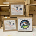 Three cardboard boxes containing Paint By Layers Craft Kits. The boxes are stacked on top of each other. The front of each box shows a circular image of the finished product. The titles of the kits are "Adventure Awaits Shadow Box," "Rocky Mountain Shadow Box," and "Under the Northern Lights Shadow Box."