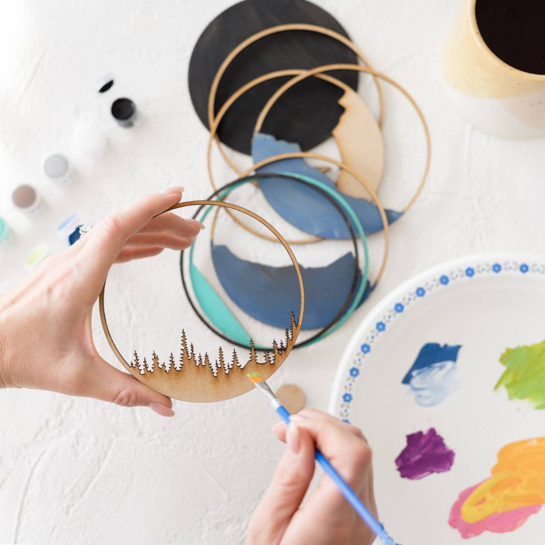 A Paint By Layers Craft Kit featuring a circular shadow box with a pre-designed landscape scene. The scene depicts a sunset over mountains, a lake, and trees. The kit includes layers of wood, paint, and instructions.