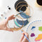 A Paint By Layers Craft Kit featuring a circular shadow box with a pre-designed landscape scene. The scene depicts a sunset over mountains, a lake, and trees. The kit includes layers of wood, paint, and instructions.