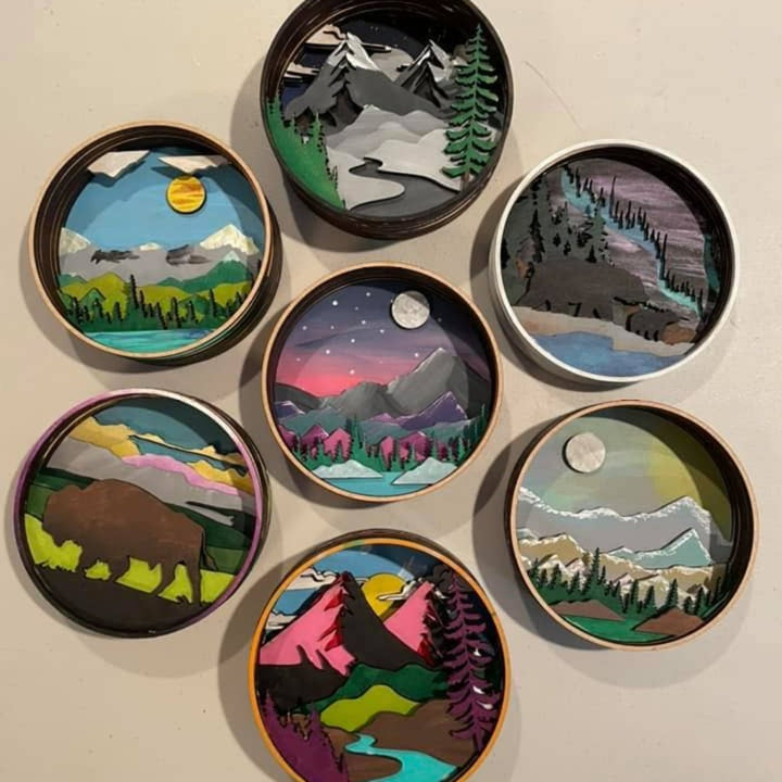 Eight circular, layered wooden shadow boxes depicting different nature scenes. The scenes include mountains, lakes, rivers, trees, a sunset, a starry night sky, a moon, and a bear. The boxes are painted in various colors and are arranged in a grid pattern on a gray surface.