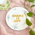 Decorative plate with 'SHAKE IT OFF' text on a pink surface with leaves